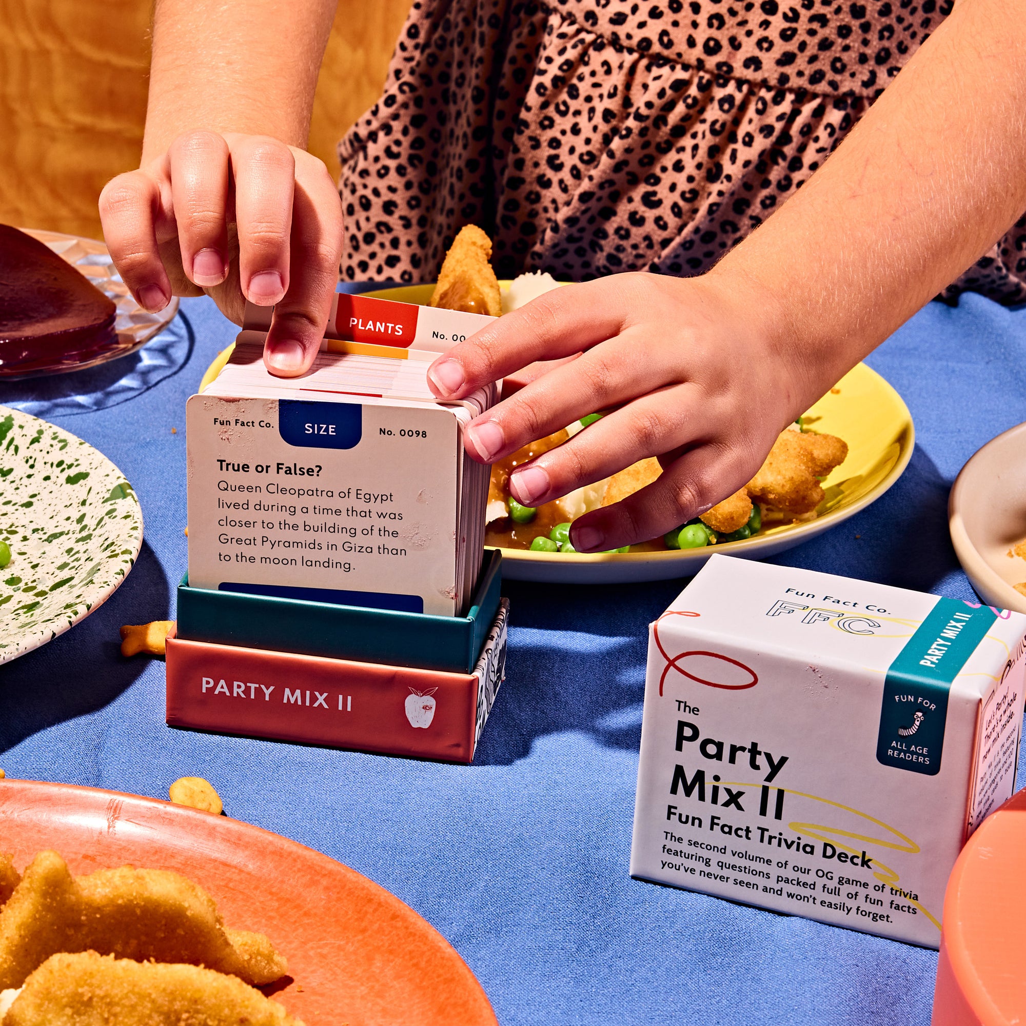 Person opening a box of 'The Party Mix II' Fun Fast Trivia Deck on a table with food.