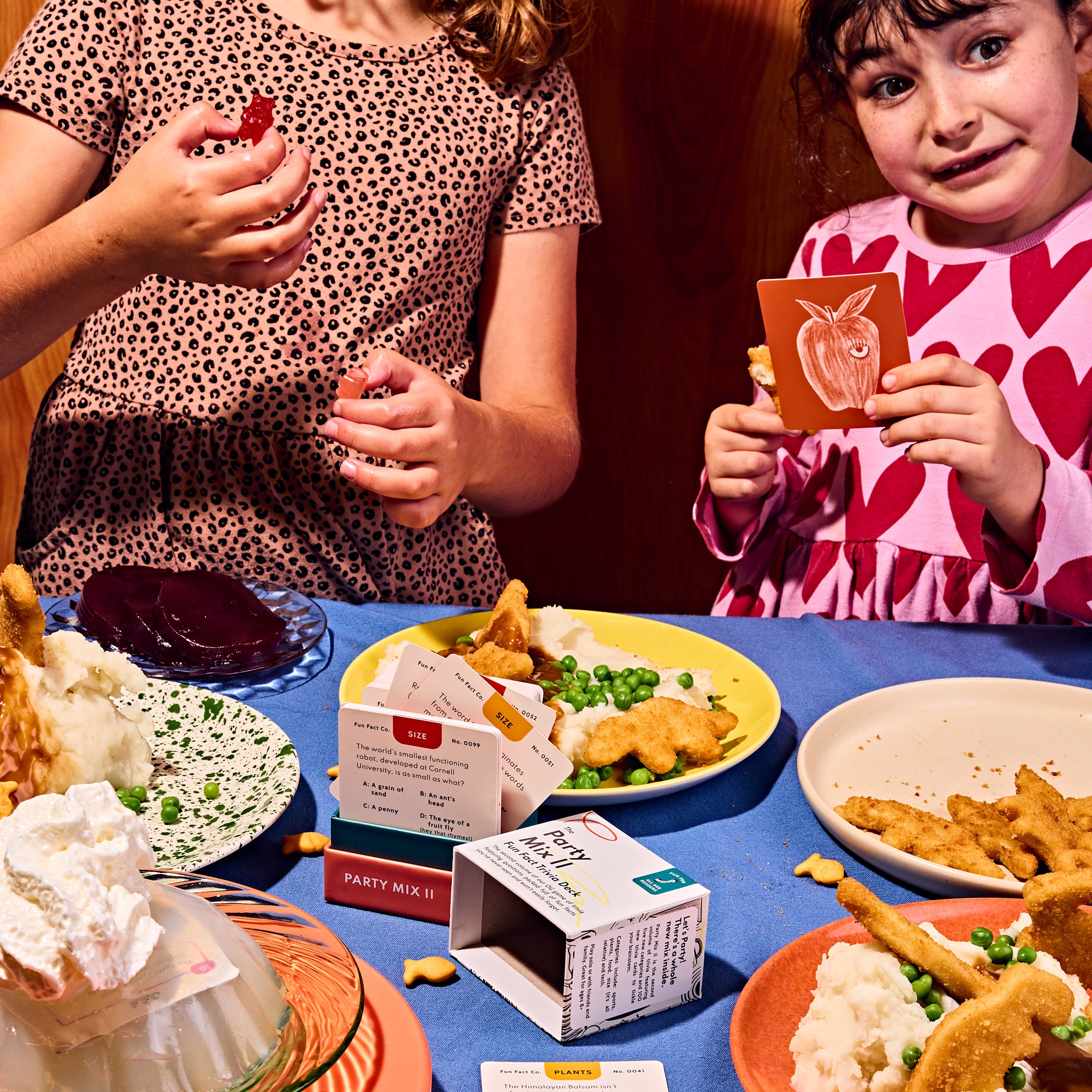 Children at a table with food and party mix boxes, one child holding a card with an apple illustration.