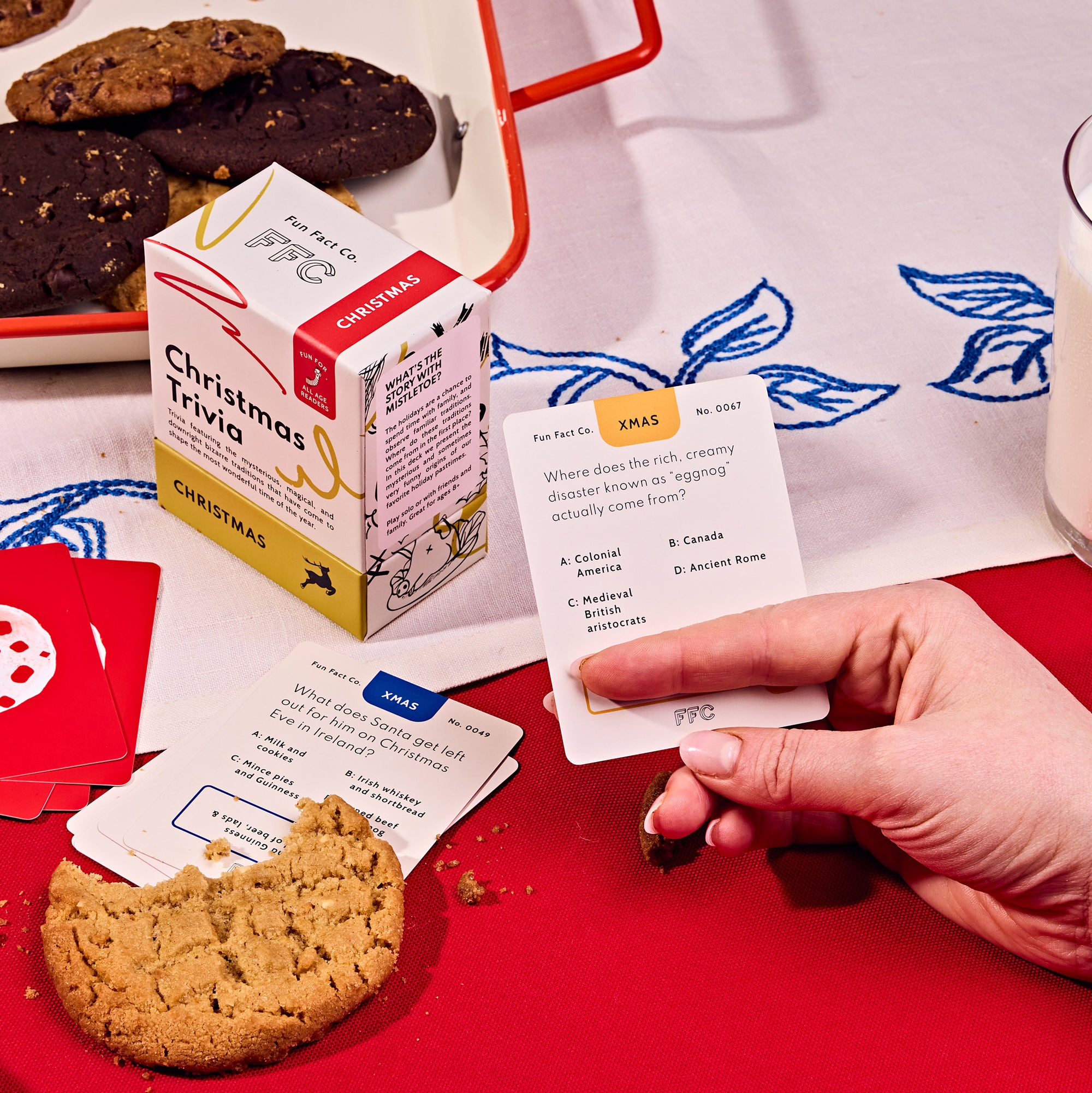 Person playing Christmas trivia with cookies and a box of trivia cards on a red table.